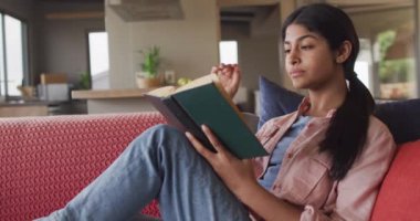 Video of biracial woman sitting on sofa and reading book. Relax, leisure and spending free time at home with technology concept.