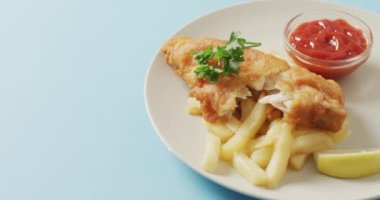 Video close up of fish and chips, parsley and bowl of ketchup on plate, with copy space on blue. tasty hot homemade fast food meal.