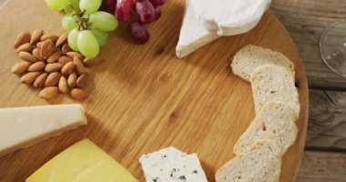 Video of cheeses, bread, nuts and grapes on board, with wine glass on wooden table. quality light food snack and drink.