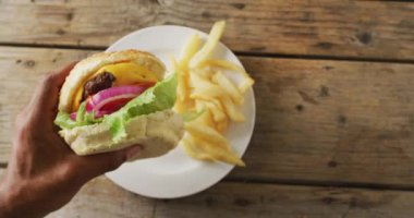Video pov of african american hand holding cheeseburger, over chips on plate on wooden table. tasty hot homemade fast food meal. ov