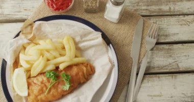Video of fish and chips on plate, salt and pepper and ketchup on wooden table. tasty hot fast food meal.