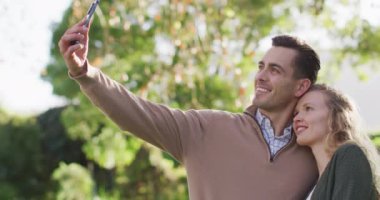 Video of happy caucasian husband and wife taking selfie smartphone in garden. Family, love and togetherness concept digitally generated video.
