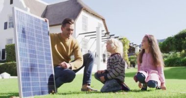 Video of happy caucasian father explaining solar panel to son and daughter in sunny garden. Family, domestic life and togetherness concept digitally generated video.