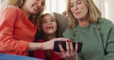 Video of happy caucasian mother, daughter and grandmother on couch looking at smartphone together. Family, domestic life and togetherness concept digitally generated video.