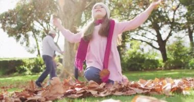 Video of happy caucasian daughter throwing autumn leaves in garden while family rakes them up behind. Family, domestic life and togetherness concept digitally generated video.