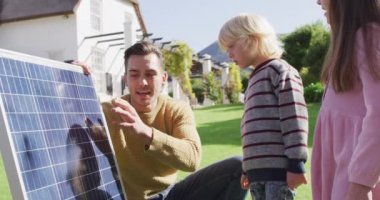 Video of happy caucasian father explaining solar panel to son and daughter in sunny garden. Family, domestic life and togetherness concept digitally generated video.