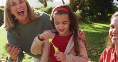 Video of smiling caucasian granddaughter blowing bubbles with mother and grandmother in garden. Family, domestic life and togetherness concept digitally generated video.