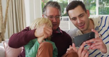 Video of happy caucasian father, grandfather and grandson sitting on couch looking at smartphone. Family, domestic life and togetherness concept digitally generated video.
