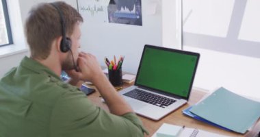 Kafkasyalı bir adam masa başında oturuyor, kulaklık takıyor ve laptopunu yavaş çekimde online toplantı için kullanıyor. Covid 19 koronavirüs karantina süresince sosyal mesafeler.