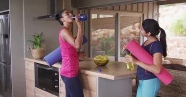 Video of happy diverse female friends holding yoga mats and talking at home. Friendship, spending quality time together at home.