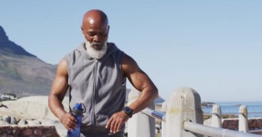 Kıdemli Afro-Amerikan adam deniz kenarındaki kayalarda içme suyu kullanıyor. Koronavirüs covid 19 salgını sırasında sağlık sağlığı için kendini izole etme emeklilik yaşam tarzı.