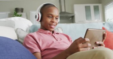 Kanepede tablet kullanan kulaklıklı mutlu Afrikalı Amerikalı çocuğun videosu. Çocukluk, ev kavramında teknolojiyle zaman geçirmek..
