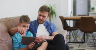 Beyaz adam ve oğlu dijital tablet kullanıyor ve gülüyorlar, evdeki kanepede ağır çekimde oturuyorlar. Kapalı alan 19 Coronavirüs karantinası süresince birlikte sosyal mesafeli zaman geçirmek..