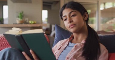 Video of biracial woman sitting on sofa and reading book. Relax, leisure and spending free time at home with technology concept.