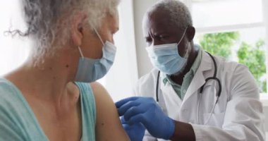 Erkek Afro-Amerikan doktor yüz maskesi takıyor ve yaşlı kadın hastaya kovid 19 aşısı enjekte ediyor. Salgın sırasında korona virüsü tedavisi ve aşı kavramı.