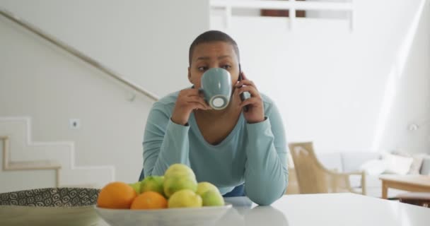 幸せなアフリカ系アメリカ人のプラスサイズの女性のコーヒーを飲んで キッチンでスマートフォンで話しています 家庭生活や余暇をテクノロジーで楽しんでいます — ストック動画