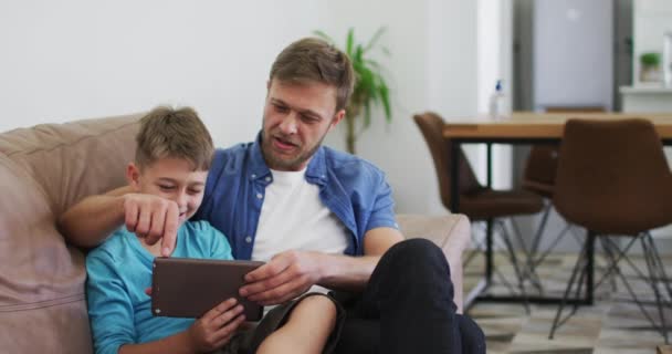 白人男性と彼の息子は ゆっくりとした動きで 自宅のソファに座って デジタルタブレットと笑いを使用しています Covid 19コロナウイルスの隔離ロックダウン中に質の高い時間を一緒に過ごす — ストック動画