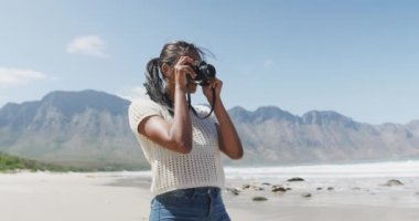 Sahilde dijital kamerayla fotoğraf çeken Afrikalı Amerikalı bir kadın. Doğa keşfi macera turizmi ve sağlıklı yaşam tarzı konsepti