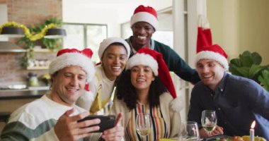 Noel Baba şapkalı bir grup mutlu arkadaş yemeği kutluyor, Noel zamanı selfie çekiyor. Yılbaşı kutlamaları, evde arkadaşlarla kutlama..