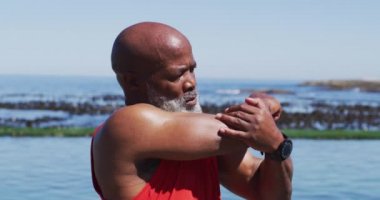 Kıdemli Afro-Amerikan adam deniz kenarındaki kayalarda egzersiz yapıyor. Koronavirüs covid 19 salgını sırasında sağlık sağlığı için kendini izole etme emeklilik yaşam tarzı.