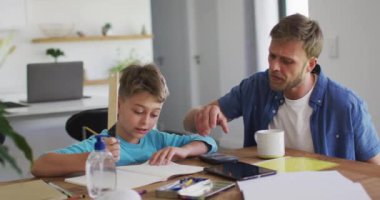 Beyaz adam oğluna ödevlerinde yardım ediyor, evde yemek masasında oturuyor, çocuk ağır çekimde dinlerken açıklama yapıyor. Kapalı alan 19 Coronavirüs karantinası süresince birlikte sosyal mesafeli zaman geçirmek..