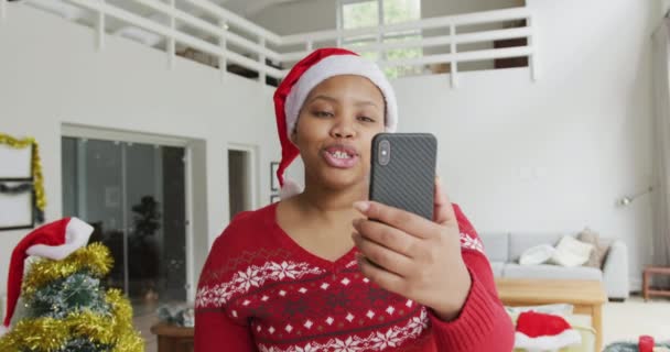 サンタの帽子の幸せなアフリカ系アメリカ人とサイズの女性は クリスマスにスマートフォンでビデオ通話を行います クリスマス冬伝統お祝い家で — ストック動画