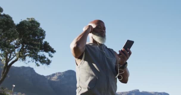シニアアフリカ系アメリカ人の男性が スマートフォンをイヤホンにして運動をしています コロナウイルス中の健康フィットネス自己分離退職生活 Covid 19パンデミック — ストック動画