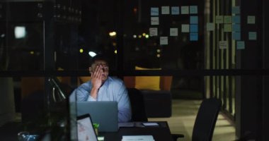 Masada dizüstü bilgisayarla oturan yorgun melez iş adamının videosu. Geceleri ofiste yalnız çalışıyor. İş, iletişim, kapsayıcılık ve dijital olarak oluşturulan esnek çalışma konsepti.