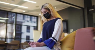 Ofisteki dinlenme odasında maske takan düşünceli beyaz iş kadınının akıllı telefon kullandığı bir video. Coronavirus konsepti sırasında dijital olarak üretilen iş, iletişim ve esnek çalışma.