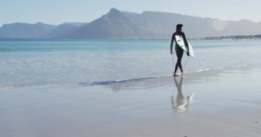 Dalgıç kıyafetli, sörf tahtası taşıyan, güneşli sahilde yürüyen beyaz bir adamın videosu. Özgürlük, spor, hobiler ve dijital olarak üretilen sağlıklı aktif yaşam tarzı konsepti.
