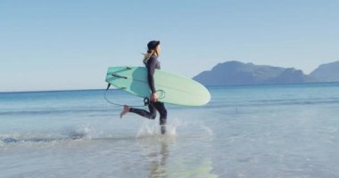 Güneşli sahilde sörf tahtası taşıyan, rasta giysili beyaz bir adamın videosu. Özgürlük, spor, hobiler ve dijital olarak üretilen sağlıklı aktif yaşam tarzı konsepti.