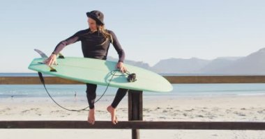 Dalgıç kıyafetli beyaz bir adamın kumsalda sörf tahtasını kontrol edişinin videosu. Özgürlük, spor, hobiler ve dijital olarak üretilen sağlıklı aktif yaşam tarzı konsepti.