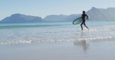Güneşli sahilde sörf tahtası taşıyan, rasta giysili beyaz bir adamın videosu. Özgürlük, spor, hobiler ve dijital olarak üretilen sağlıklı aktif yaşam tarzı konsepti.