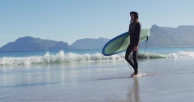 Dalgıç kıyafetli, sörf tahtası taşıyan, güneşli sahilde yürüyen beyaz bir adamın videosu. Özgürlük, spor, hobiler ve dijital olarak üretilen sağlıklı aktif yaşam tarzı konsepti.