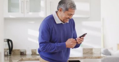 Mutfakta akıllı telefon kullanan, gülümseyen ve gülen üst düzey melez bir adamın videosu. Emeklilik, iletişim, kapsayıcılık ve kıdemli yaşam tarzı konsepti.