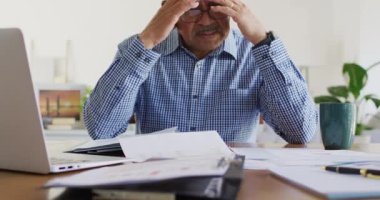 Masada oturan, evrak işlerine bakan ve evde dizüstü bilgisayar kullanan yaşlı, melez bir adamın videosu. İş iletişimi, uzaktan çalışma, kapsayıcılık ve kıdemli yaşam tarzı konsepti.