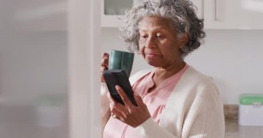 Akıllı telefon kullanan ve mutfakta kahve içen son sınıf öğrencisi Afro-Amerikan kadının portresi. Emeklilik ve ev kavramında zaman geçirme.