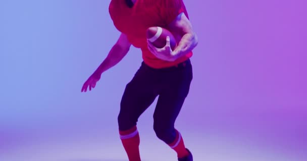 ネオンの紫色の背景にボールとヘルメットで白人アメリカ人サッカー選手のビデオ アメリカンフットボール スポーツ 競技のコンセプト — ストック動画
