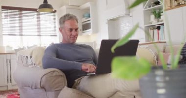 Oturma odasındaki kanepede dizüstü bilgisayar kullanarak oturan mutlu beyaz adam. Evde yalnız vakit geçirmek..