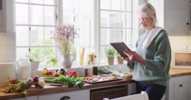 Mutfakta durup tablet kullanarak yemek hazırlayan son sınıf beyaz kadın. Evde yalnız zaman geçirmek..