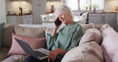 Oturma odasındaki koltukta oturan, dizüstü bilgisayar kullanan ve akıllı telefondan konuşan son sınıf beyaz kadın. Evde yalnız zaman geçirmek..