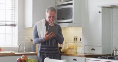 Önlük giyen mutlu beyaz adam, mutfakta duruyor, yemek pişiriyor ve tablet kullanıyor. Evde yalnız vakit geçirmek..