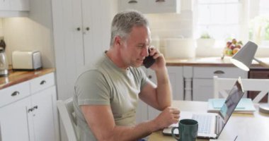 Mutfakta masada oturan, dizüstü bilgisayar kullanan ve akıllı telefondan konuşan mutlu beyaz adam. Evde yalnız vakit geçirmek..