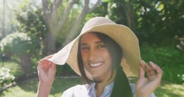 Güneşli bahçede güneş şapkası takıp kameraya gülümseyen mutlu melez kadının video portresi. Mutluluk, sağlık, ev hayatı ve kapsayıcılık kavramı.