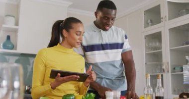 Mutfakta birlikte yemek hazırlayan mutlu çiftin tablet tarifini kullandıkları bir video. İletişim, mutluluk, sağlık, ev hayatı ve kapsayıcılık kavramı.