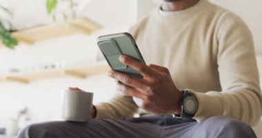 Afrika kökenli Amerikalı bir adam kahve içiyor ve akıllı telefon kullanıyor. Evde yalnız zaman geçirmek..
