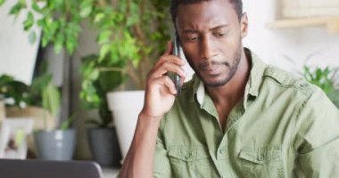 Mutlu Afro-Amerikan adam mutfakta masada oturmuş, akıllı telefondan konuşuyor. Evde yalnız zaman geçirmek..
