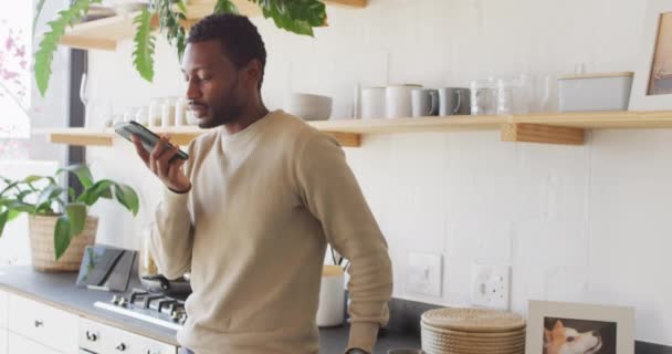 幸せなアフリカ系アメリカ人の男性キッチンでカウンターにもたれて スマートフォンで話しています 自宅だけで質の高い時間を過ごす — ストック動画