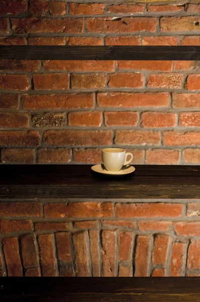Hrnek na kávu na dřevěné polici proti cihlové zdi — Stock fotografie