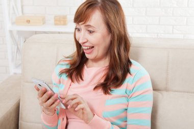 Şok olmuş, orta yaşlı, ağzı açık bir şekilde cep telefonuna bakıp evdeki kanepede oturan bir kadın. Boşluk ve yerel kavramı kopyala.